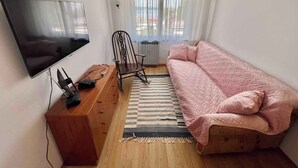 Une petite pièce bien éclairée, avec un téléviseur à écran plat fixé au mur, un bureau en bois avec une chaise, un canapé rose matelassé et un tapis à rayures.