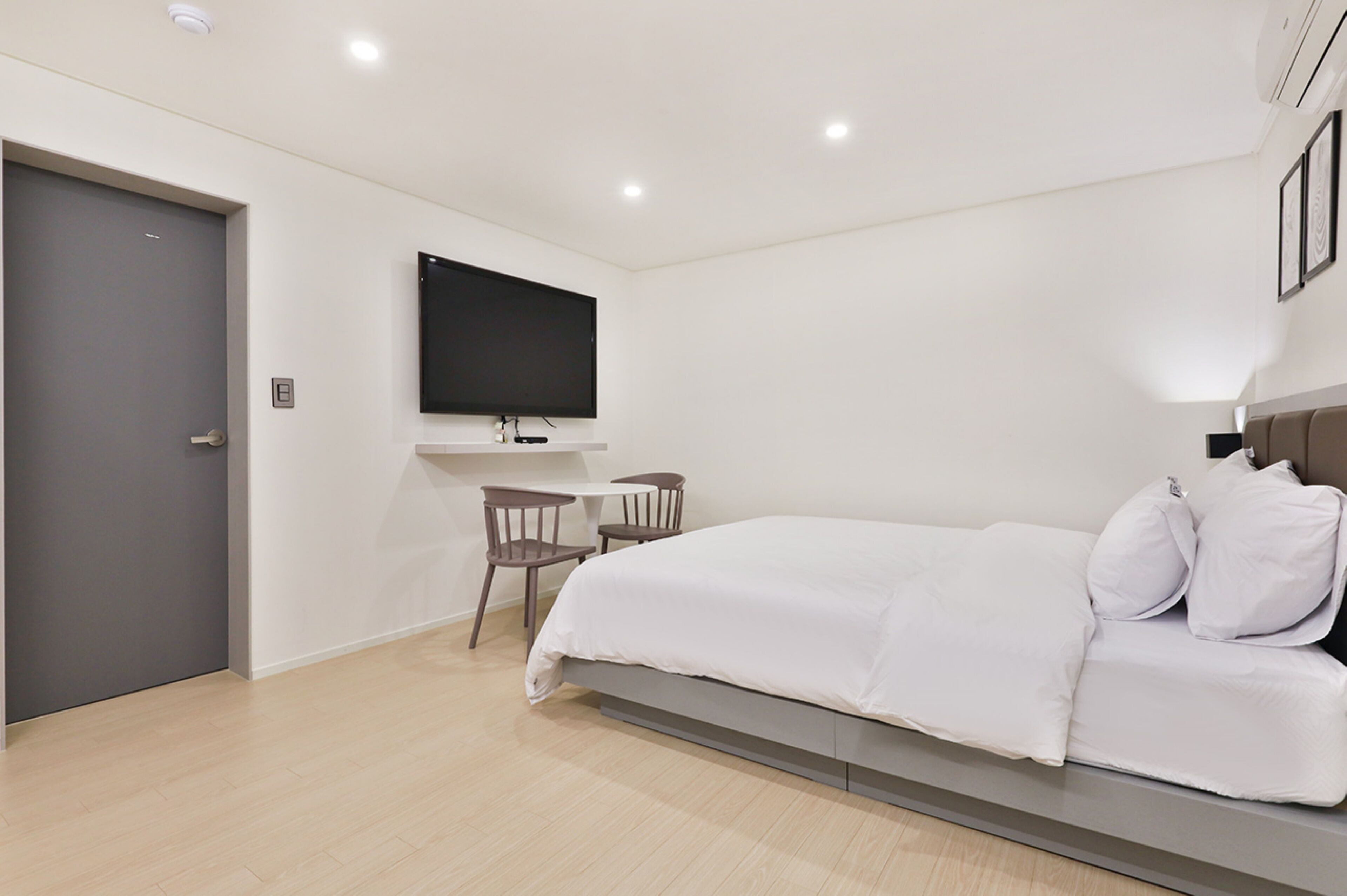 A modern hotel room with a bed, a desk, two chairs, a flat-screen TV, and a door.