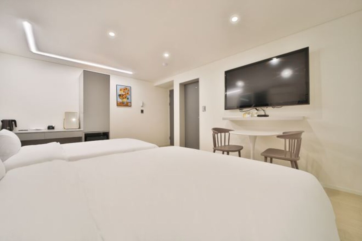 A modern hotel room with a large bed, a TV mounted on the wall, a small table with two chairs, and a desk with a chair.