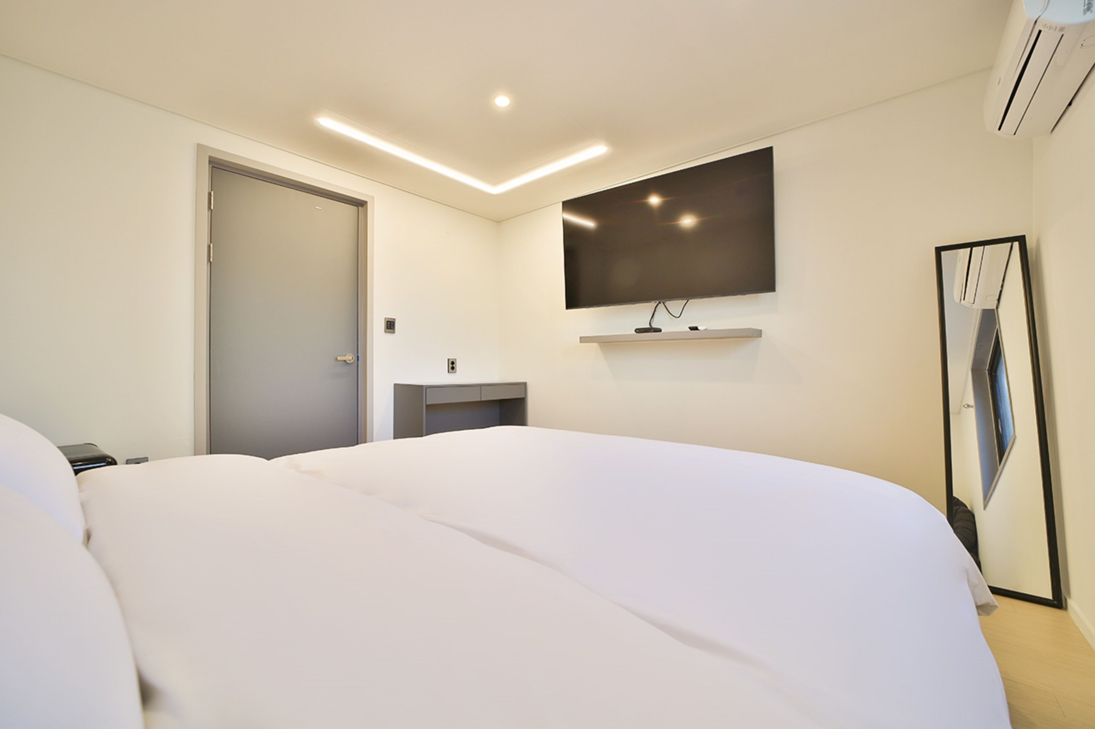 A modern hotel room with a large bed, a flat-screen TV mounted on the wall, a bedside table, and a door.