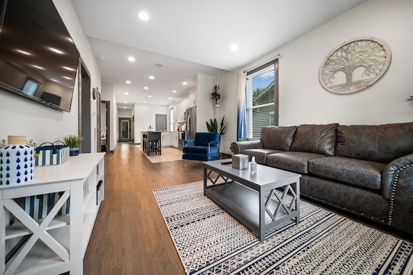 A modern living room with a brown leather sofa, a white coffee table, and a patterned rug. A kitchen is visible in the background with white cabinets and a blue chair.