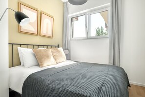 Un dormitorio ordenado con una cama, una lámpara de noche, una ventana con cortinas y dos cuadros en la pared.