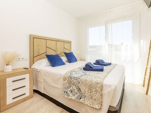A hotel room with a wooden headboard, a bedside table with drawers, a patterned throw blanket, and a view of the city through large windows.