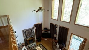 A spacious living room with a wooden ceiling, a ceiling fan, a fireplace, a sofa, a coffee table, and large windows. - Mountain Memory Cabin (Topton)