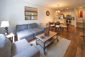A living room with a grey sofa, a coffee table, and a lamp. There is a kitchen in the background with a microwave and a refrigerator.
