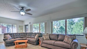 Amplia sala de estar con sofá modular, una mesa de centro de madera y un ventilador de techo. Ventanas grandes ofrecen luz natural y vistas al exterior.