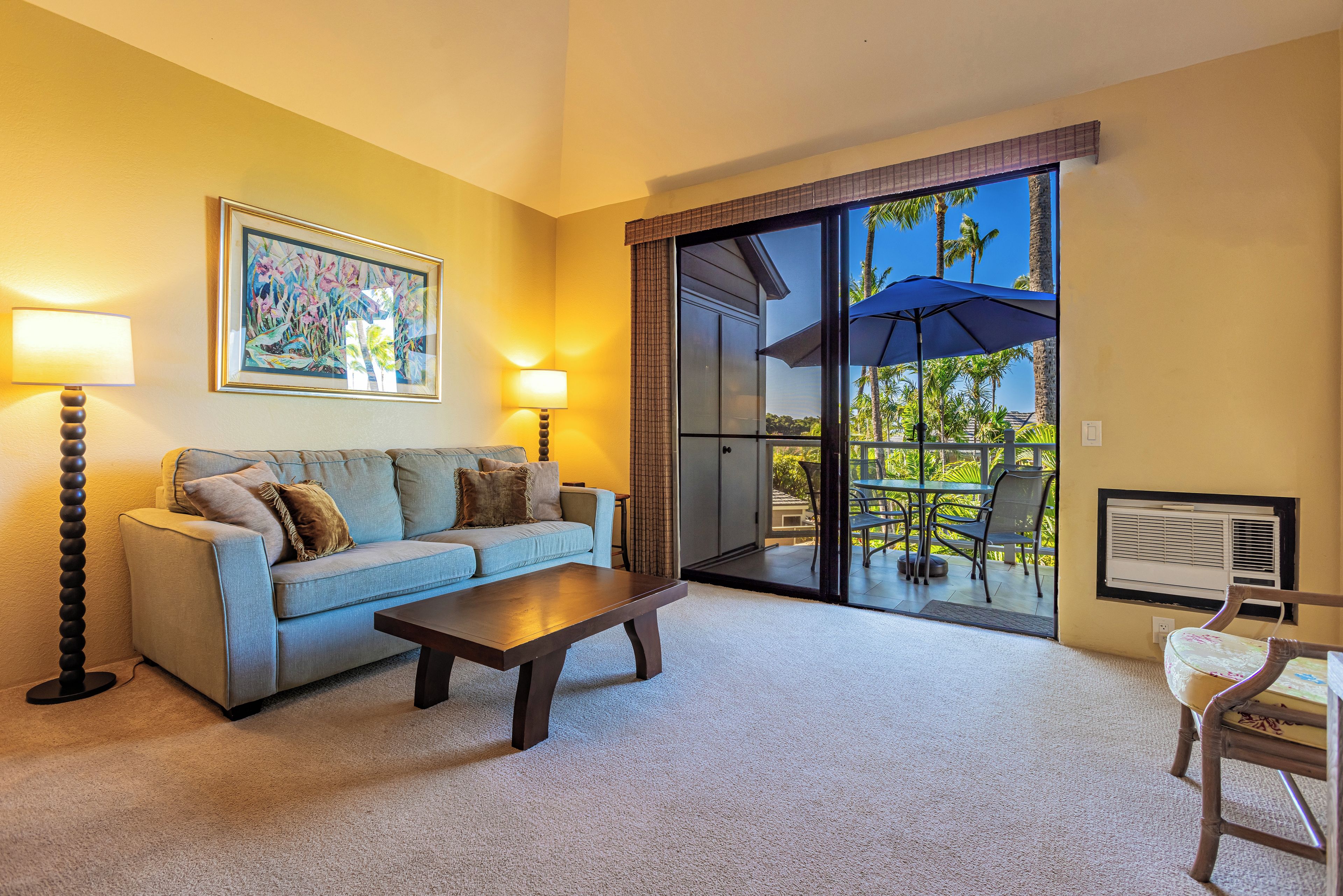 A living room with a sofa, coffee table, and two lamps. There is a sliding glass door leading to an outdoor area with chairs and an umbrella.