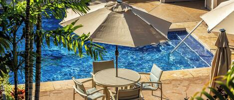 Een zwembadgedeelte met een ronde tafel en stoelen, omringd door parasols en palmbomen.