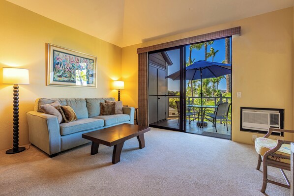 A living room with a sofa, coffee table, and two lamps. There is a sliding glass door leading to an outdoor area with chairs and an umbrella.
