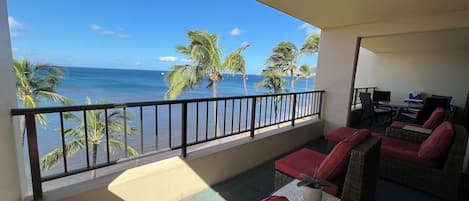 A balcony with wicker furniture, a small table, and a view of the ocean and palm trees.