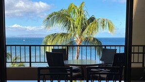 A balcony with a table and chairs, overlooking a palm tree and the ocean.