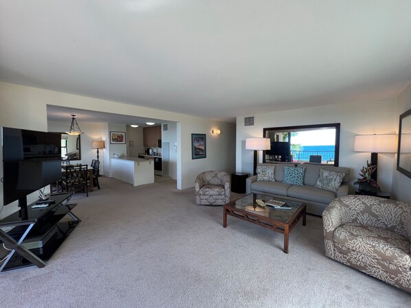 A spacious living room with a sofa, armchairs, a coffee table, and a TV. A kitchen is visible in the background, and there is a balcony with a view.