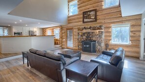 A log cabin interior with a stone fireplace, leather sofas, a dining area, and a kitchen.