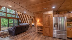 A cozy cabin interior with a wooden ceiling, a sofa, a refrigerator, and a large window offering a view of the outdoors.