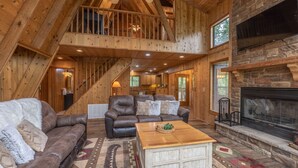 A spacious living area with a high ceiling, wooden beams, a fireplace, and a staircase leading to an upper level.