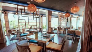 En restaurant med træborde og stole, hængende fletlamper og store vinduer med udsigt til grønne områder.