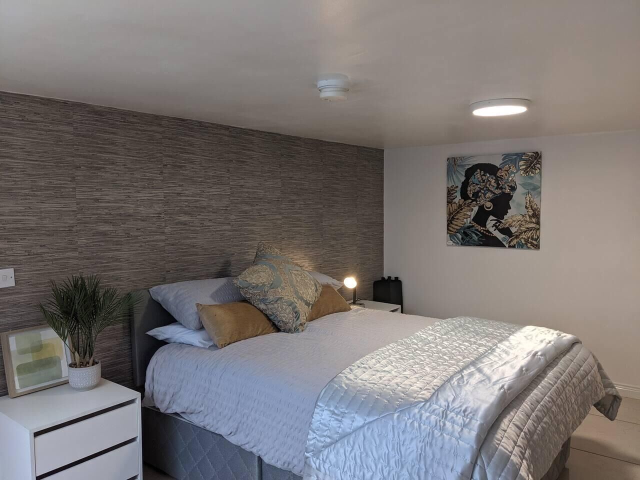 A modern bedroom with a bed, nightstands, a wall-mounted light, a framed picture, and a potted plant.