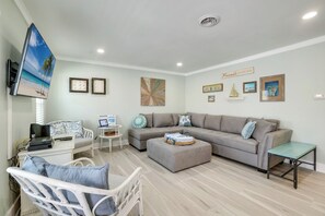 A living room with a sofa, ottoman, white armchair, small table, and a flat-screen TV mounted on the wall.
