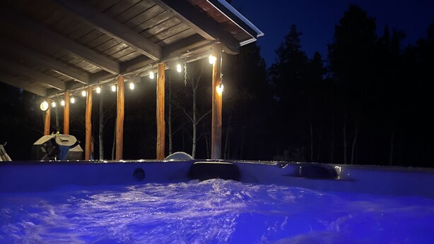 A hot tub under a wooden pergola with string lights, surrounded by snow.