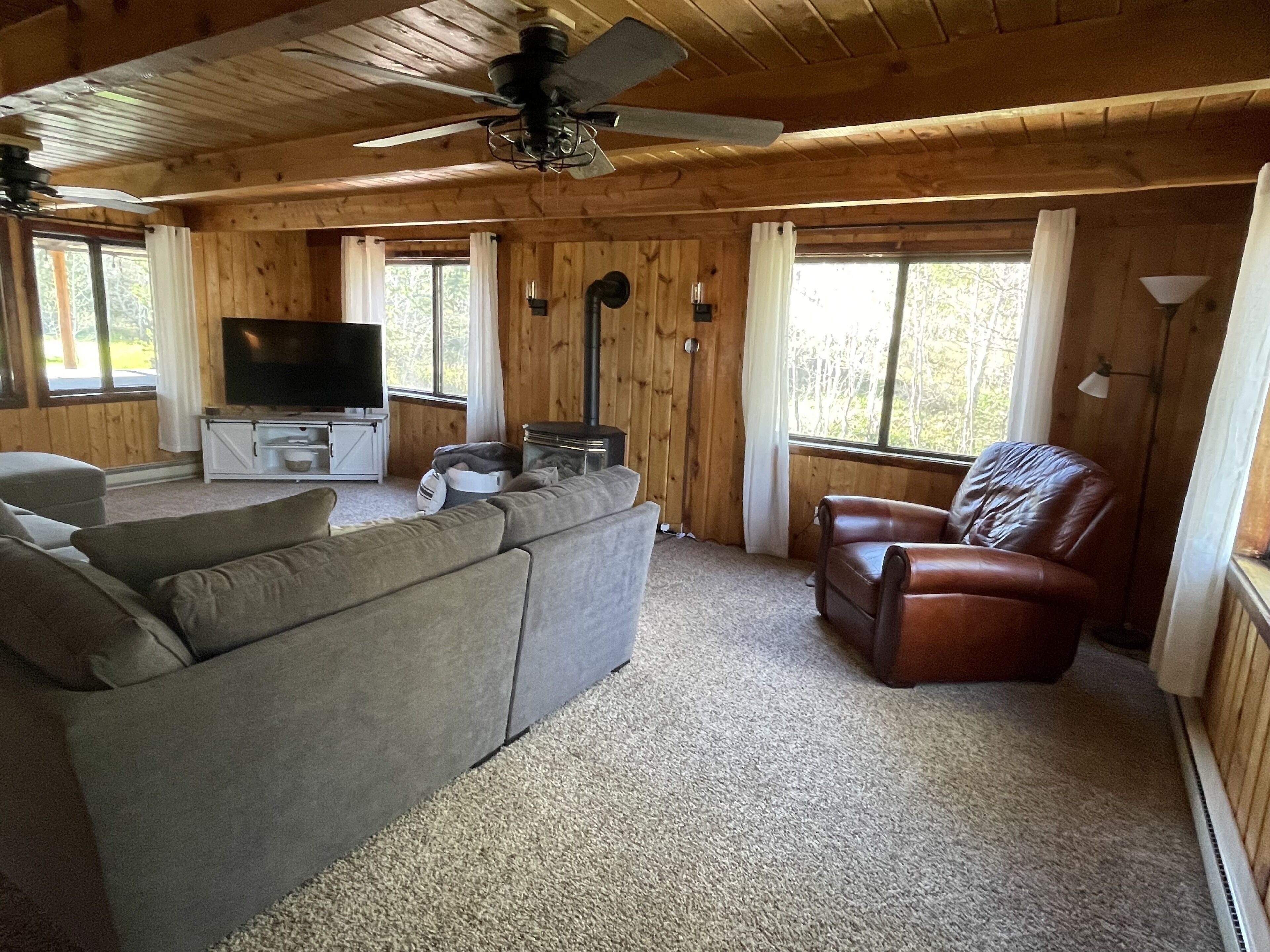 Een knus interieur van een chalet met een grijze bank, een leren fauteuil, een televisie en een houtkachel.