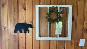 A wooden wall with a black silhouette of a bear, a white-framed window with a wreath, and a light switch.