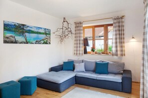 A modern living room with a sectional sofa, a painting of a lake, and a window with curtains. - Zugspitzblick Apartment with Mountain Views, Garden, and Wi-Fi (Grainau)