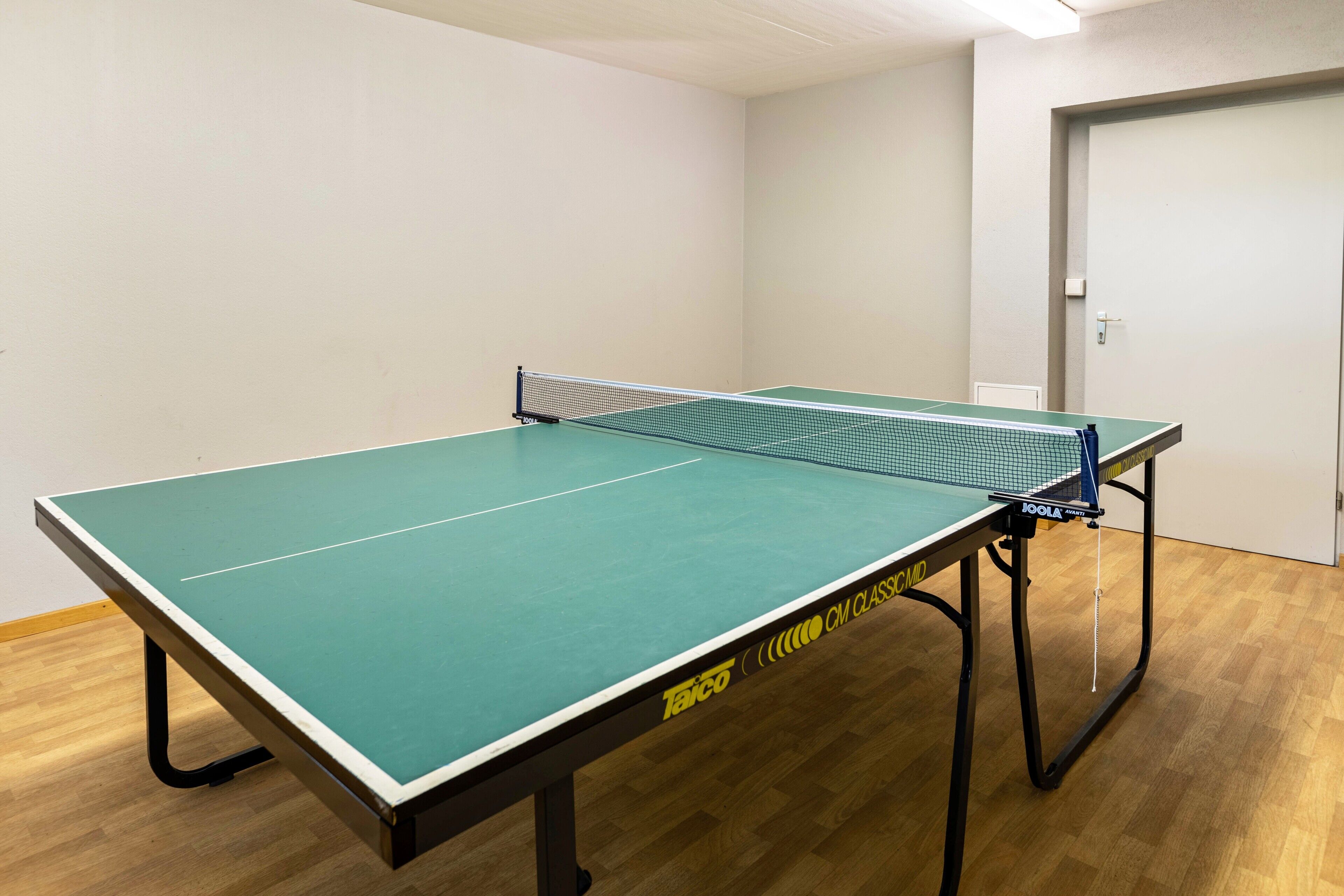 Une table de ping-pong avec un filet, dans une pièce dont le sol est en bois et où l’on aperçoit une porte au fond.