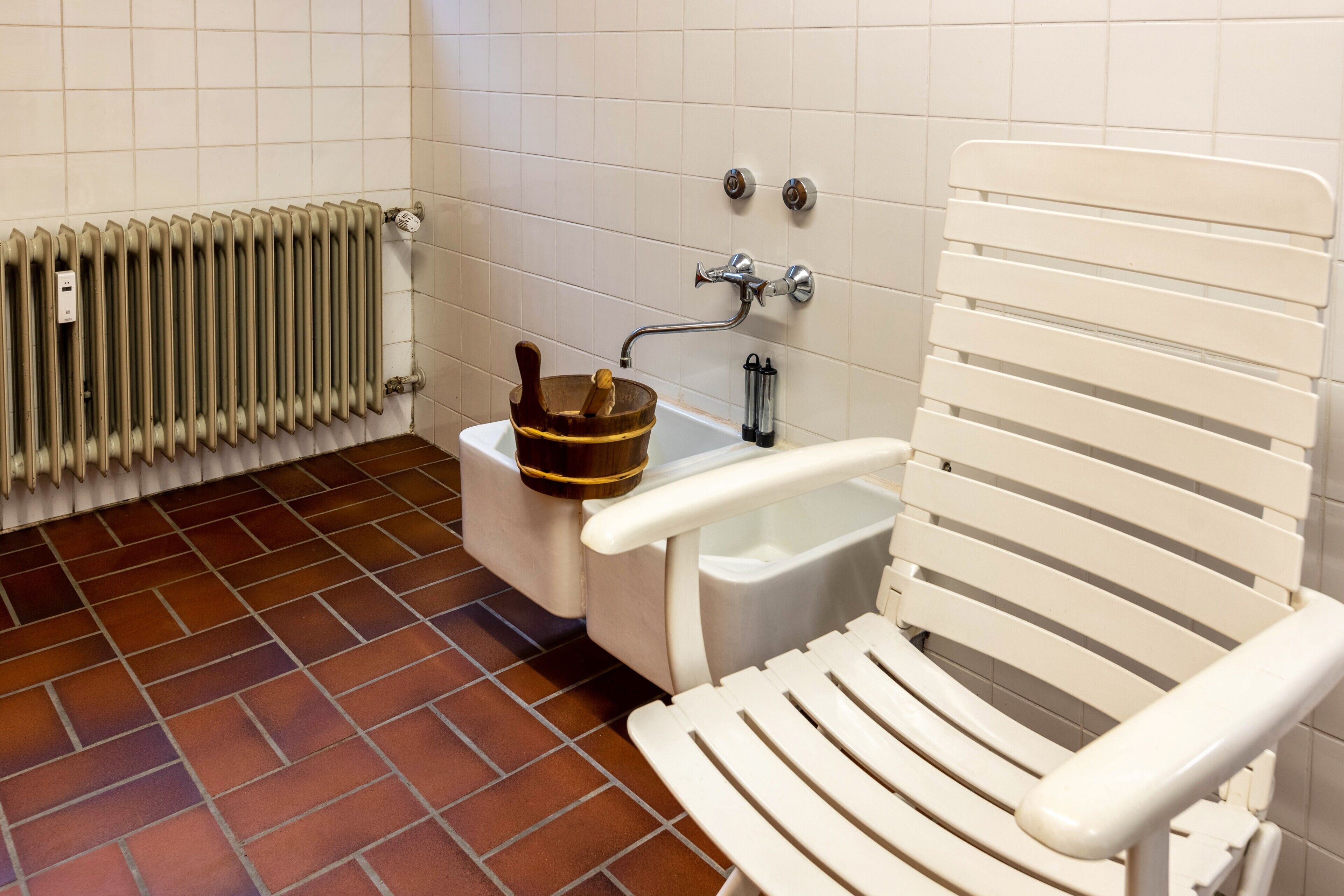 Een badkamer met een radiator, een houten emmer, een witte stoel en een wit wastafel.