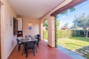 Um alpendre coberto com mesa e cadeiras, uma prateleira pequena com objetos, e vista para um jardim e árvores.