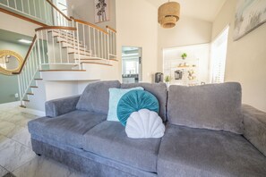 A grey sofa with teal and white cushions in a living room with a staircase and kitchen in the background. - Family Oasis: Pool & Spa! (Moreno Valley)