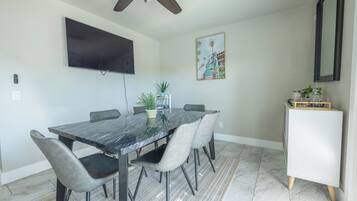 A dining area with a marble table, grey upholstered chairs, a ceiling fan, a flat-screen TV, a potted plant, and a small cabinet.