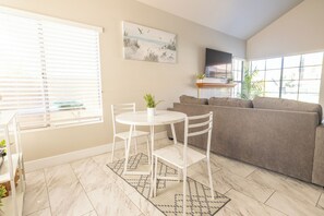 A living room with a beige sofa, a white dining table with chairs, a TV mounted on the wall, and a window with blinds.