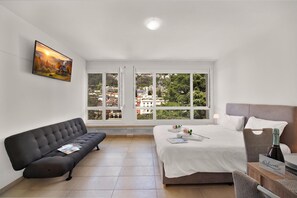 A hotel room with a bed, a sofa, a TV, and large windows offering a city view.