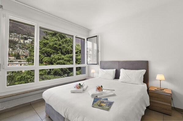 Una camera d'albergo con un'ampia finestra che si affaccia su una cittadina collinare, un letto rifatto con cuscini, un comodino con una lampada e una rivista sul letto.