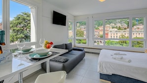 A modern hotel room with a sofa, a bed, a dining area with a table and chairs, and large windows offering a city view. - Casa Cupido Locarno Walk to Lake & Centre, Locarno, Switzerland (Locarno)