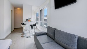 A modern living room with a grey sofa, a small dining table with two chairs, a TV mounted on the wall, and large windows. - Casa Cupido Locarno Walk to Lake & Centre, Locarno, Switzerland (Locarno)