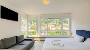 A modern hotel room with a bed, a sofa, large windows, and a view of a cityscape. - Casa Cupido Locarno Walk to Lake & Centre, Locarno, Switzerland (Locarno)