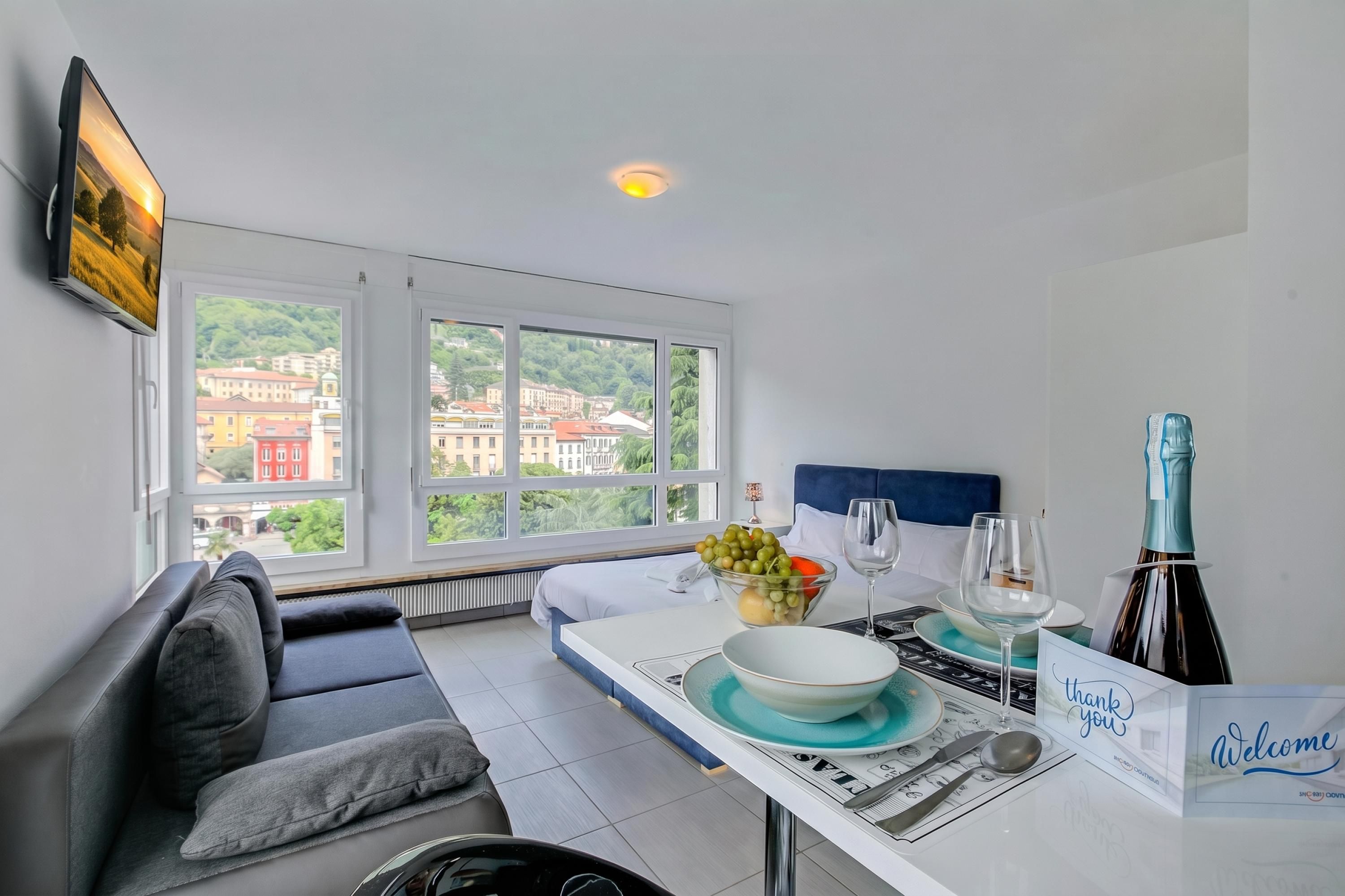 A modern hotel room with a sofa, dining table with a fruit bowl, wine glass, and a bottle.