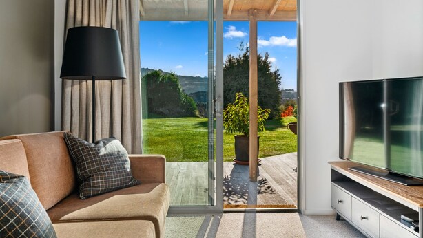 A modern living room with a sofa, a TV, and a sliding glass door leading to a garden. - Mountain View Bliss - Albert Town Holiday Unit (Albert Town)