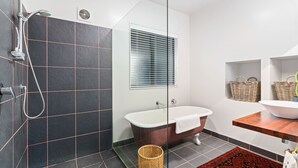 A modern bathroom with a glass shower enclosure, a freestanding bathtub, a window with blinds, wicker baskets on shelves, and a red patterned rug. - Mountain View Bliss - Albert Town Holiday Unit (Albert Town)