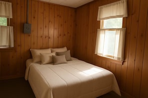 Una habitación con paneles de madera, una cama, dos ventanas con cortinas y un interruptor de luz en la pared.