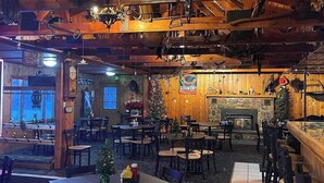 Un intérieur rustique avec des poutres en bois, une cheminée en pierre et un sapin de Noël. Des tables et des chaises sont disposées pour les repas.