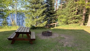 Una mesa de picnic y un pozo de fuego en un área con césped, árboles y una masa de agua al fondo.
