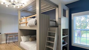 Una habitación con una cama litera, un escritorio con taburetes, una escalera y una ventana con persianas.