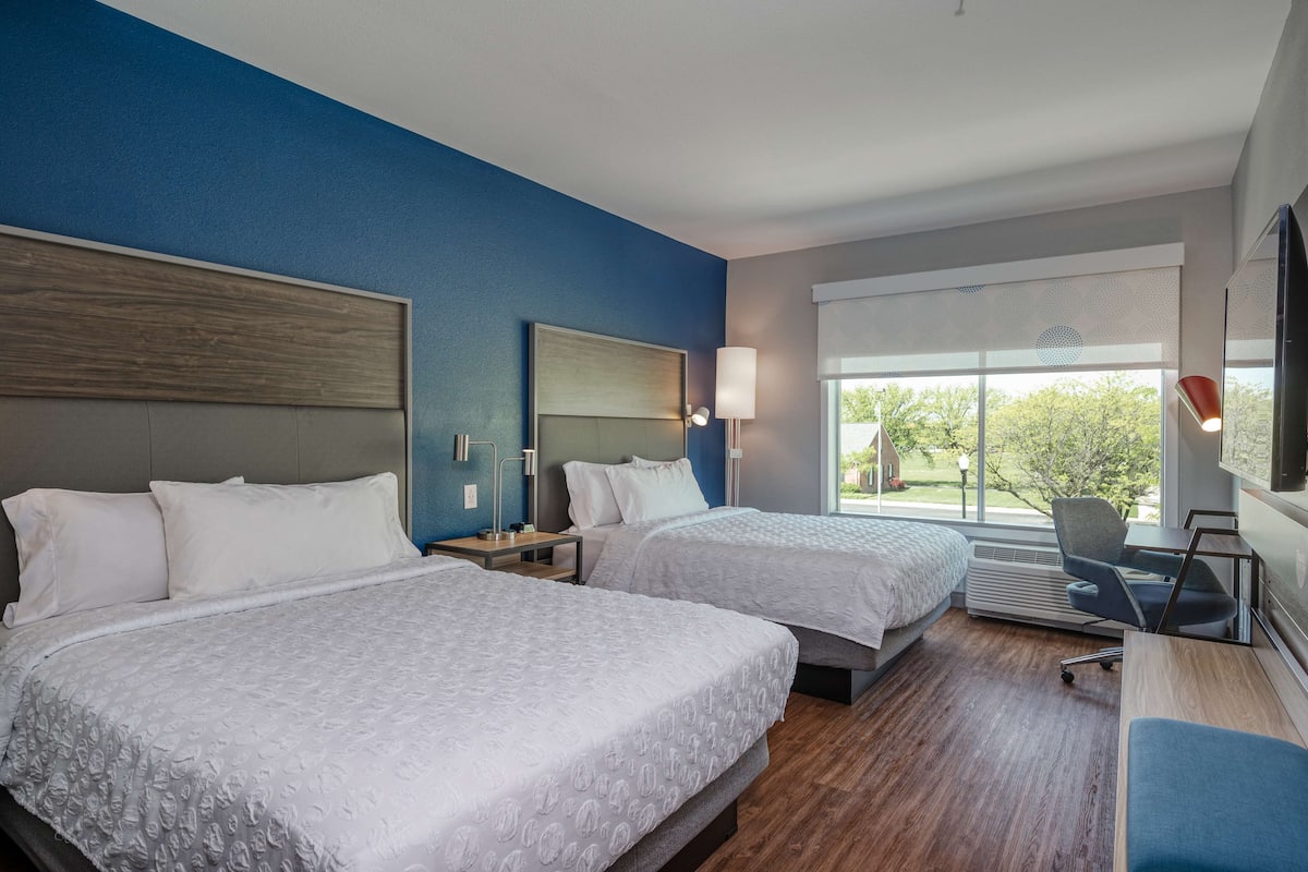 Une chambre d’hôtel avec deux lits, un bureau, une chaise et une grande fenêtre donnant sur les arbres.