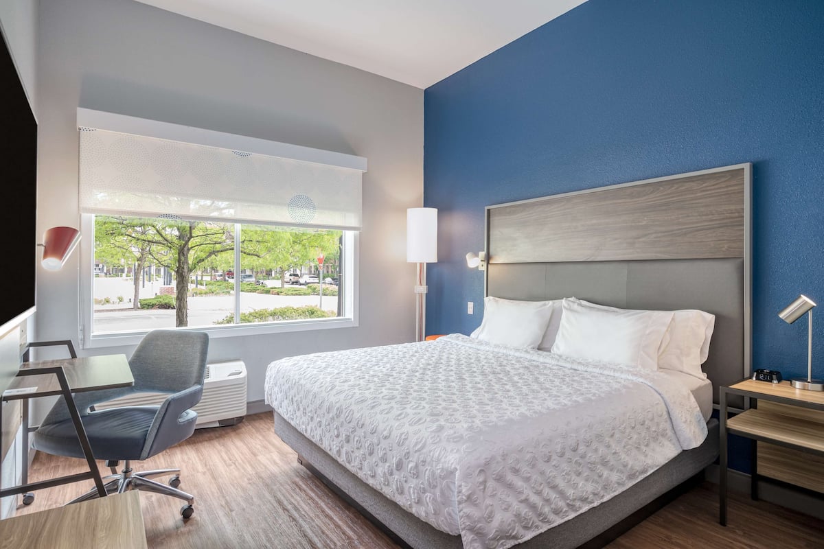 Une chambre d’hôtel avec un grand lit, un bureau et une chaise. Il y a une fenêtre donnant sur les arbres et les bâtiments à l’extérieur.