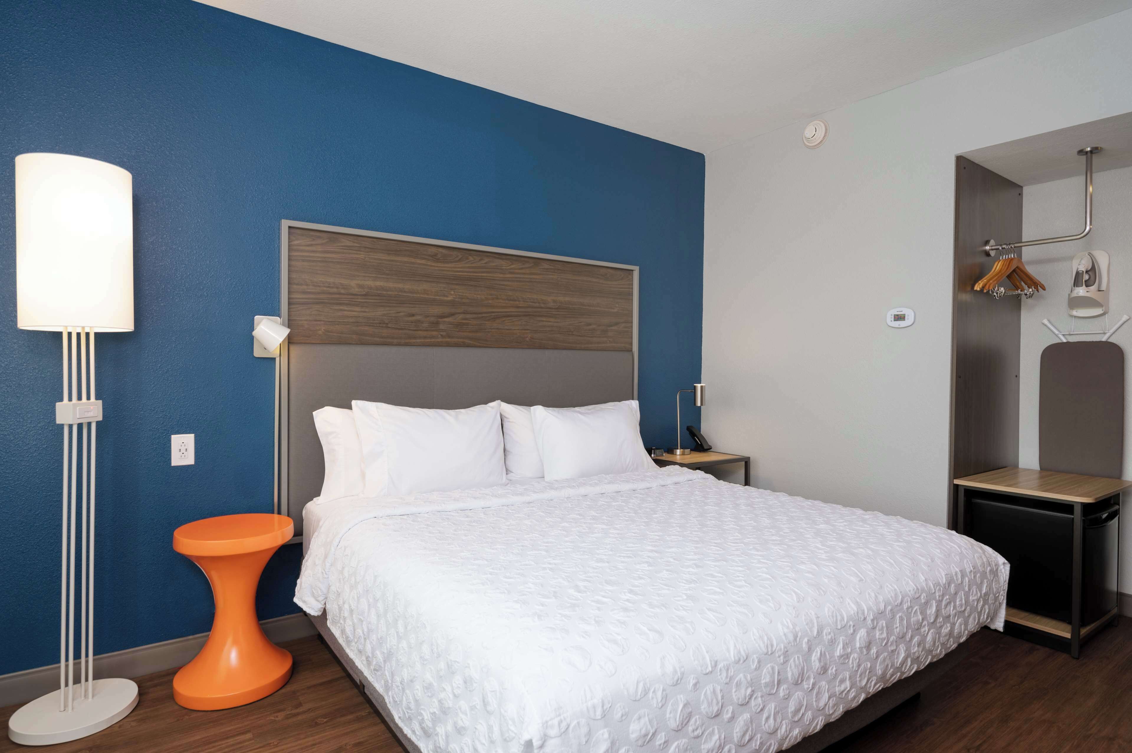 A neatly made bed with a white duvet and pillows, a modern bedside table, a floor lamp, and a small orange stool.