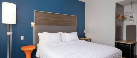 A neatly made bed with a white duvet and pillows, a modern bedside table, a floor lamp, and a small orange stool.