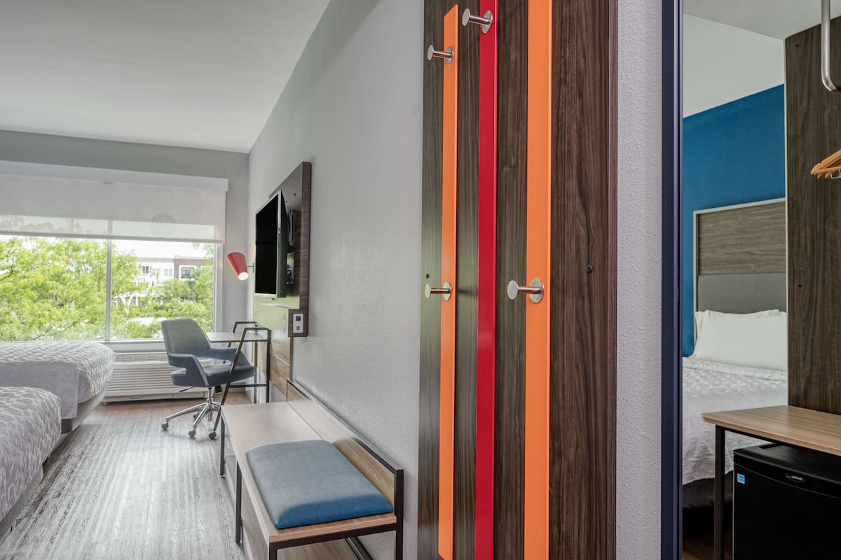 Une chambre d’hôtel moderne avec un grand lit, un bureau, une chaise et une petite table. Un téléviseur est fixé au mur et une fenêtre offre une vue sur les arbres à l’extérieur.