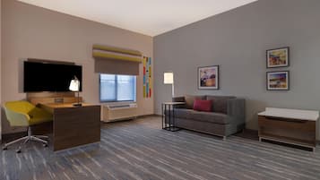 A hotel room with a flat-screen TV, a desk with a chair, a sofa, a standing lamp, and a small table.